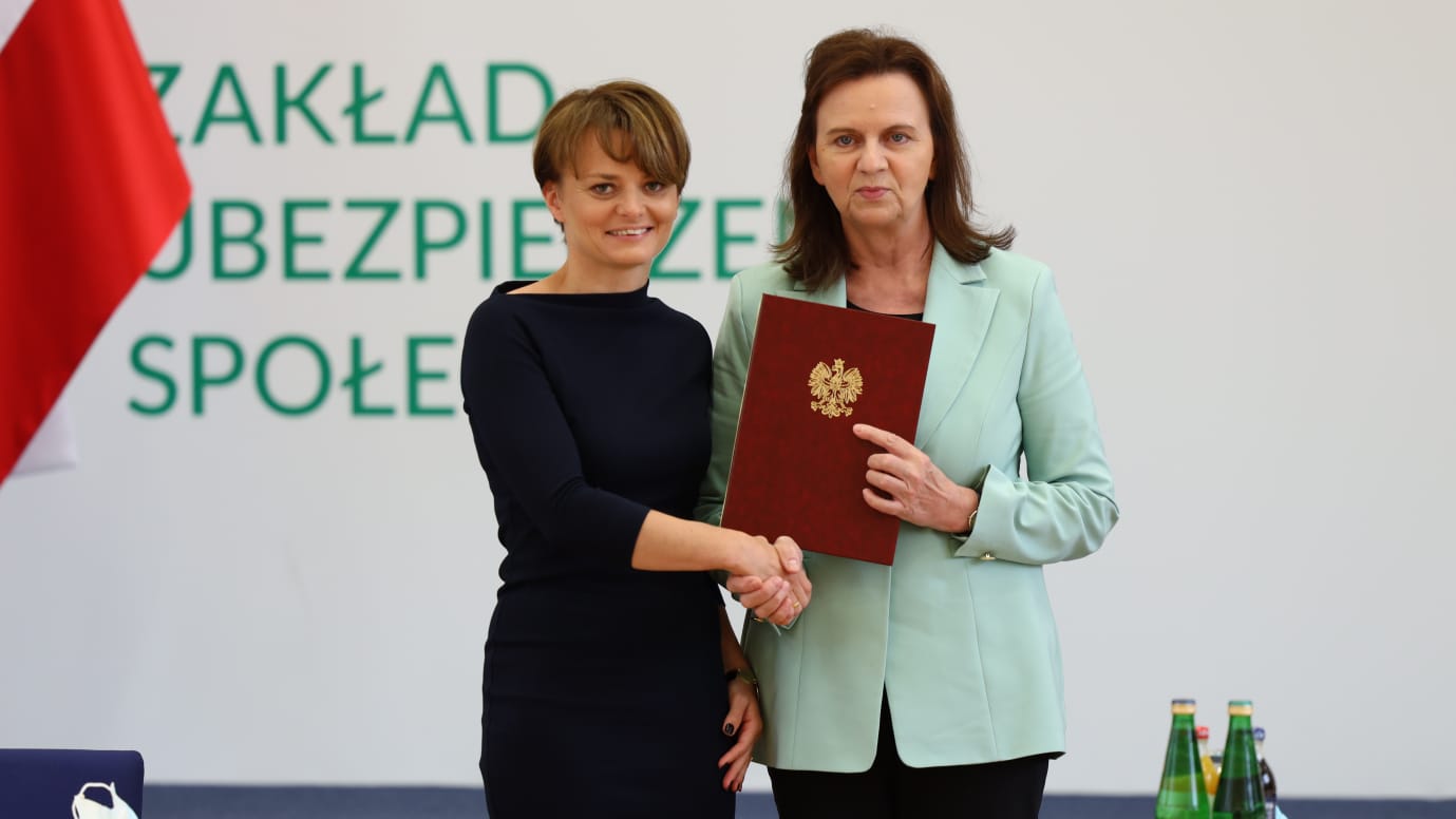 Minister Jadwiga Emilewicz i prezes Gertruda Uścińska stoją na białym tle z napisem Zakąłd ubęzpieczeń społecznych, podają sobie ręce, prezez Uścińska trzyma bordową teczkę ze złotym orłem, w rogu lewym fragment flagi, czerwony